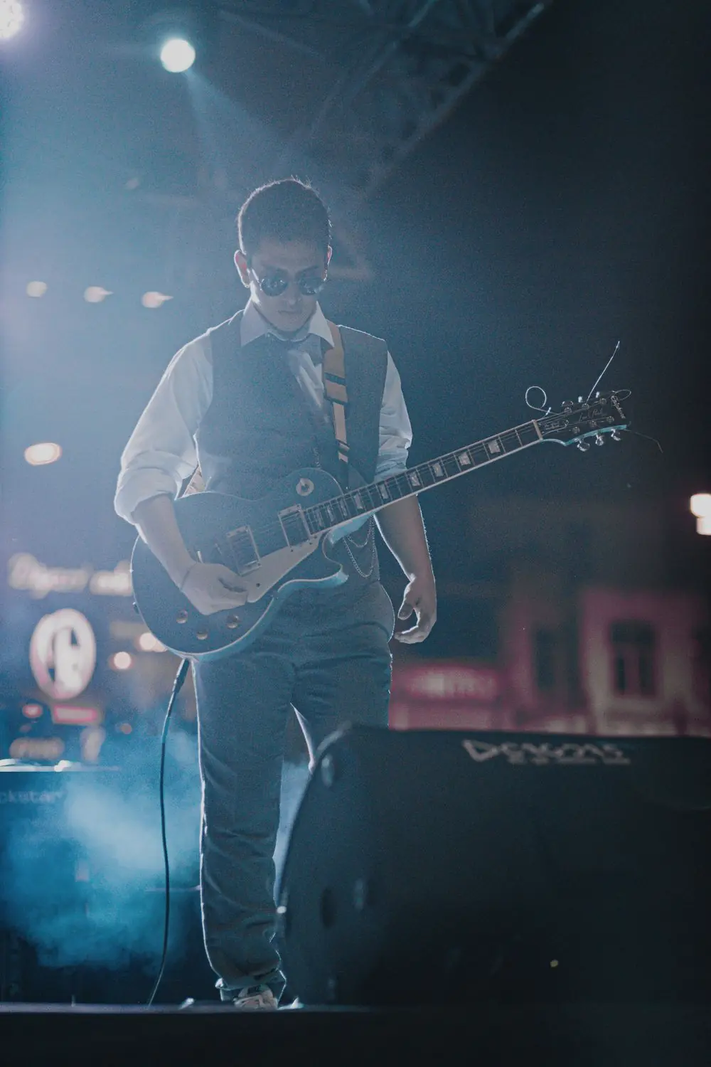Mehmet playing a dark electric guitar under cool blue stage lights at an outdoor show.
