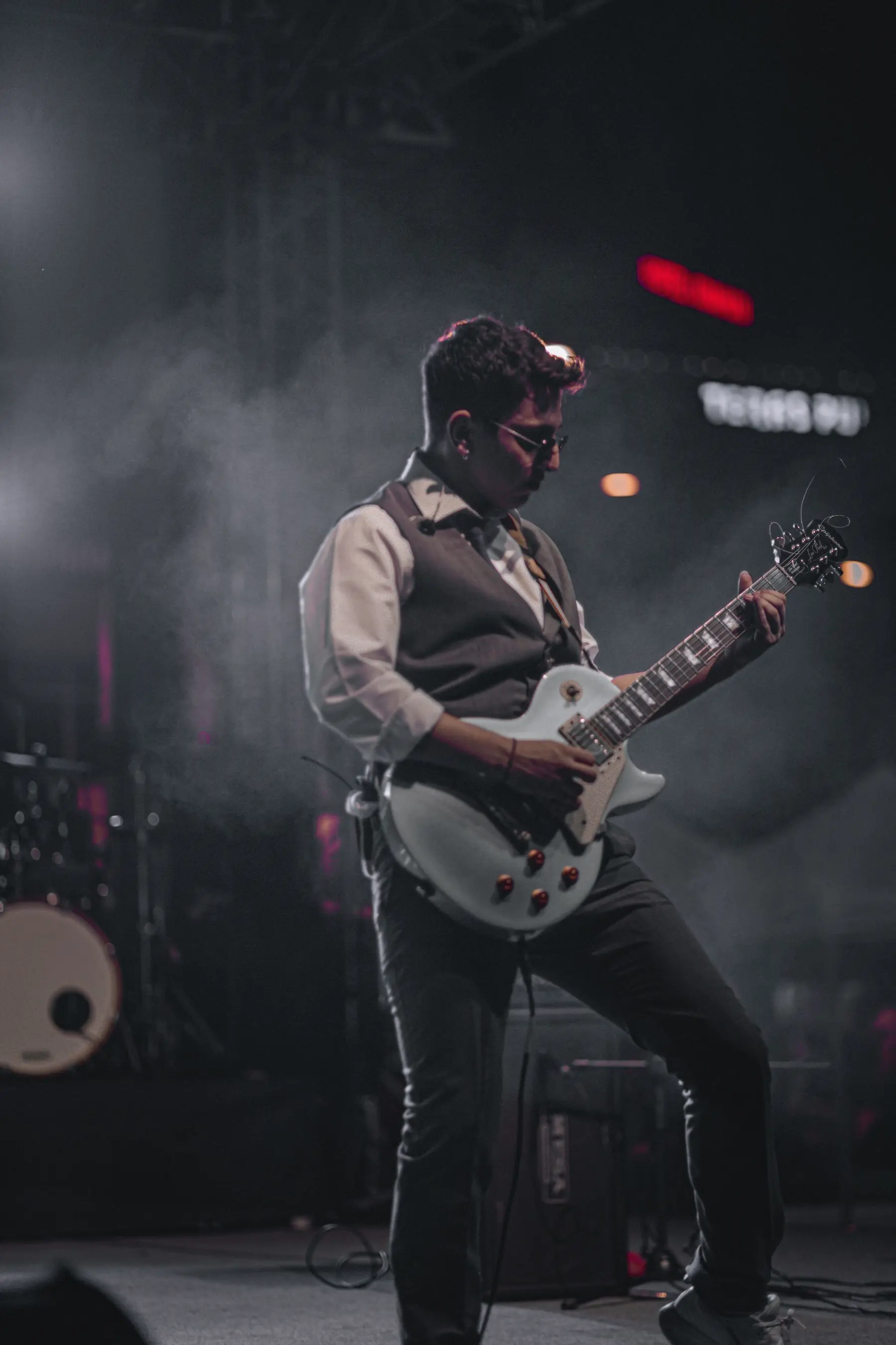 Mehmet playing a white electric guitar on stage through stage smoke, lit in red.
